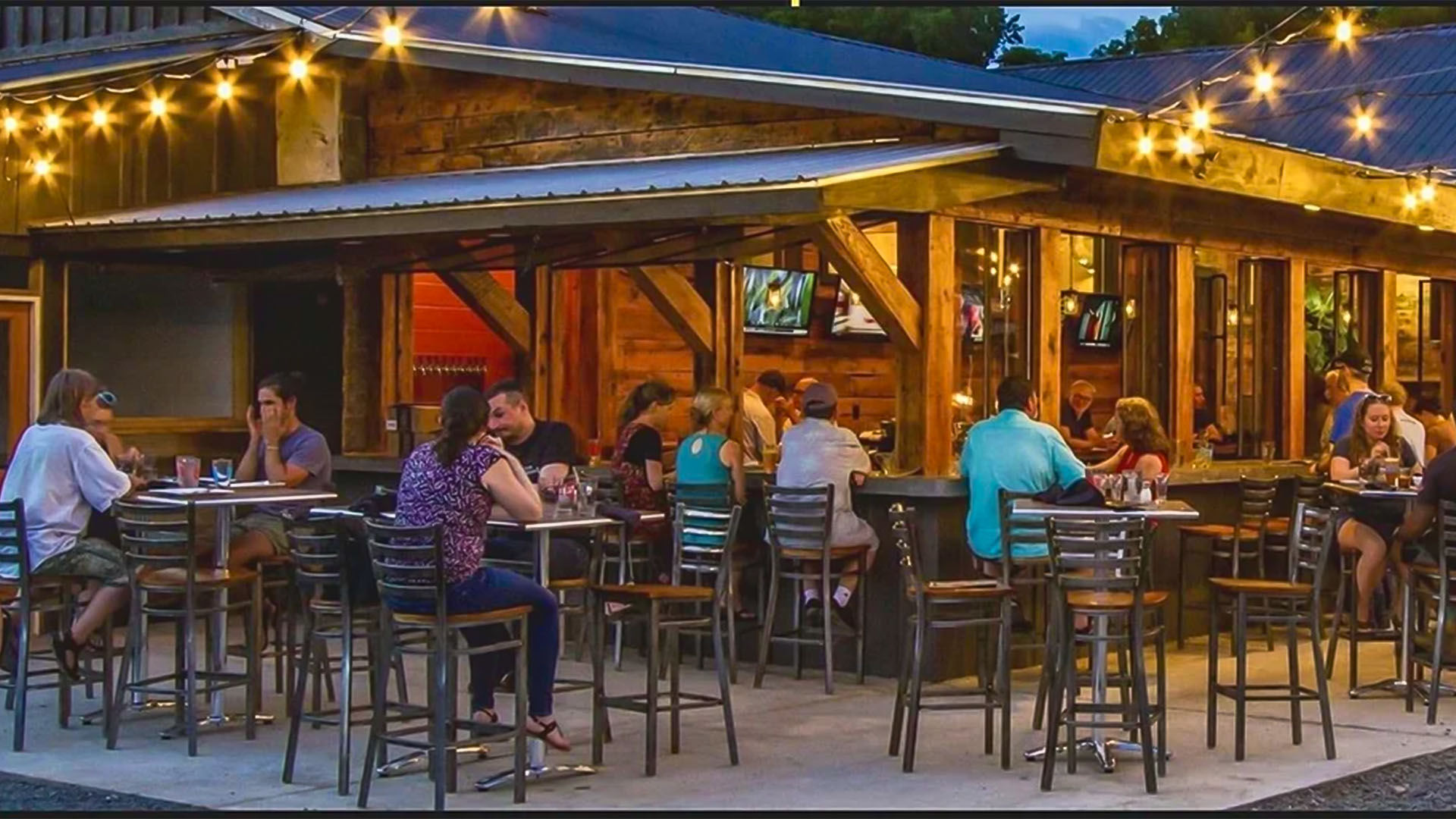 People sit at outdoor tables, eating and drinking, under warm string lights at a rustic wooden restaurant patio.