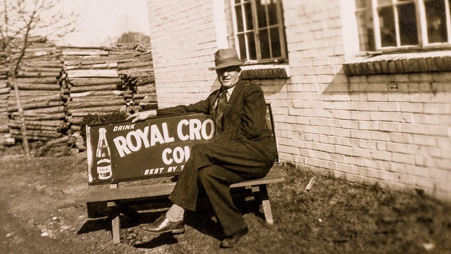 Man in suit and hat sitting on a bench with a Royal Crown Cola sign beside a brick building and wood pile.