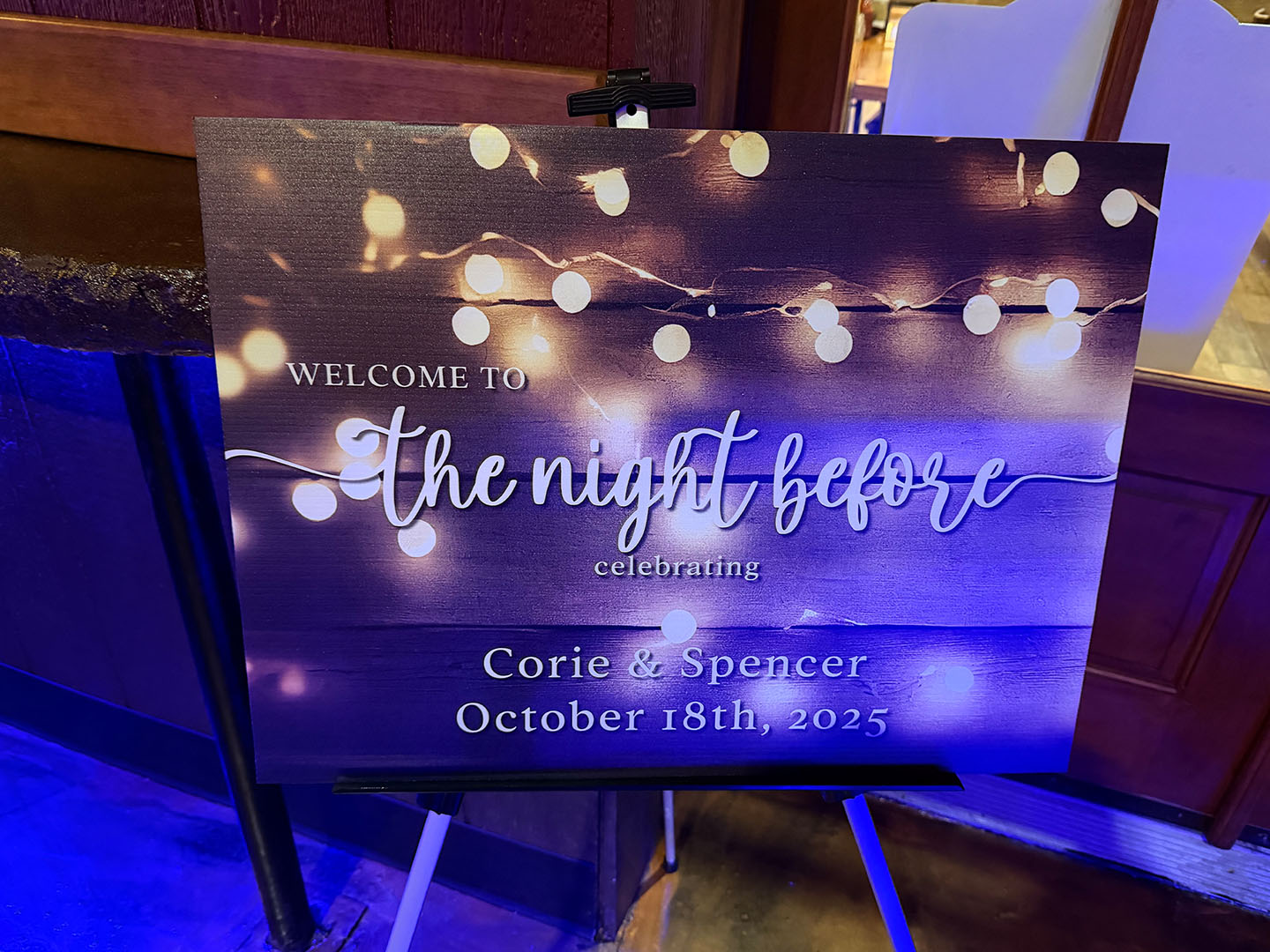 A sign with string lights reads: Welcome to the night before, Corie & Spencer, October 18th, 2025.