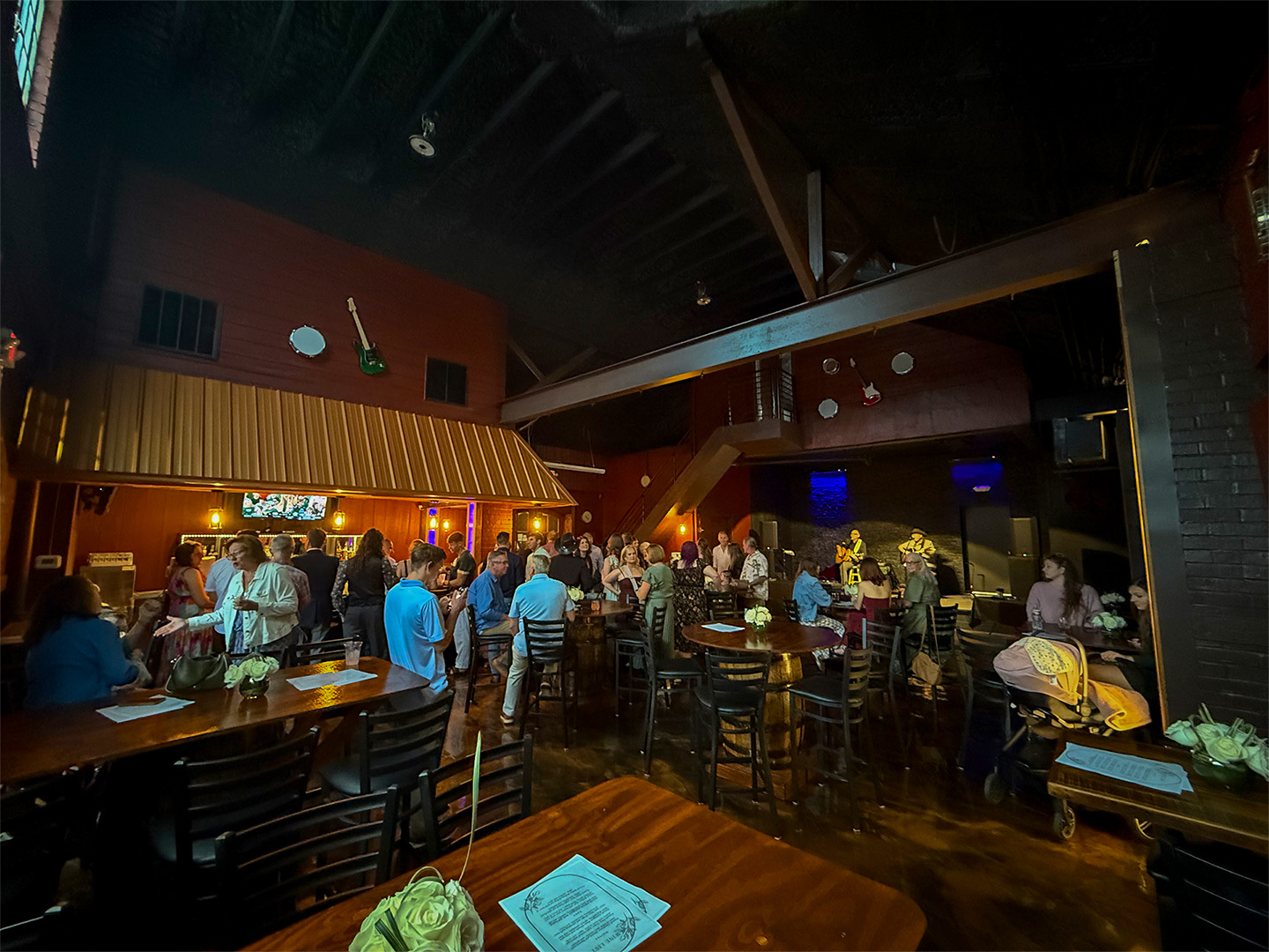 People gather in a dimly lit bar with wooden tables, chatting and standing near a small stage area.